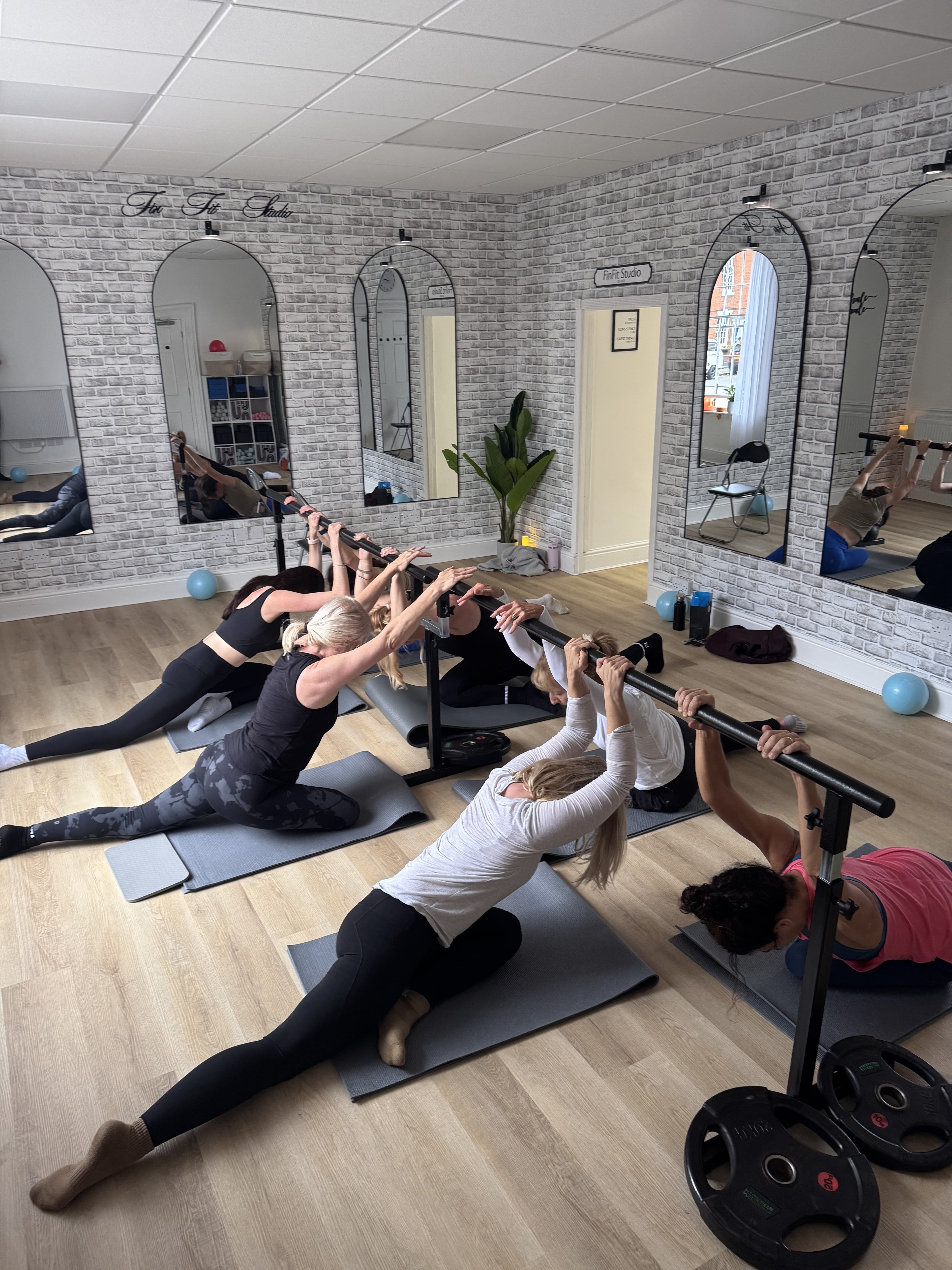 Six women in a deep kneeling side-stretch reaching to the barre